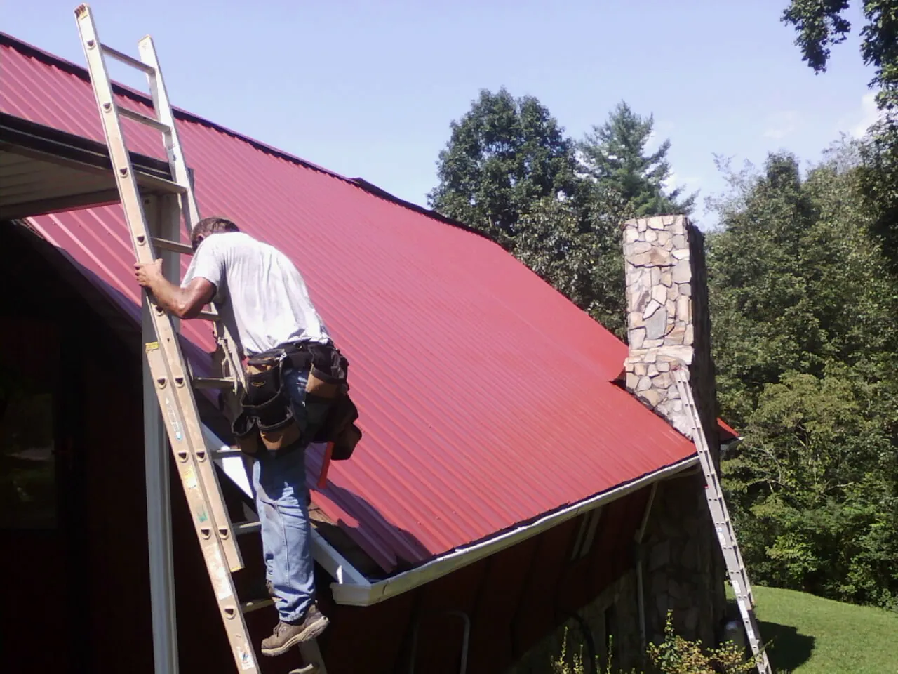 Roofer on a ladder beside a red metal roof and stone chimney — Soffit & Fascia Repair by Roofing Brothers in Jesup